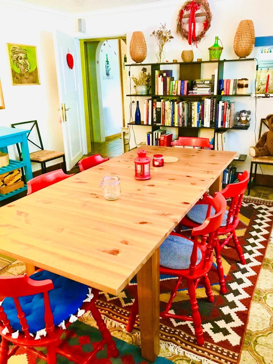 Gemütlicher Aufenthaltsraum im Haus Feuchtl mit großem Holztisch, roten Stühlen und Bücherregal